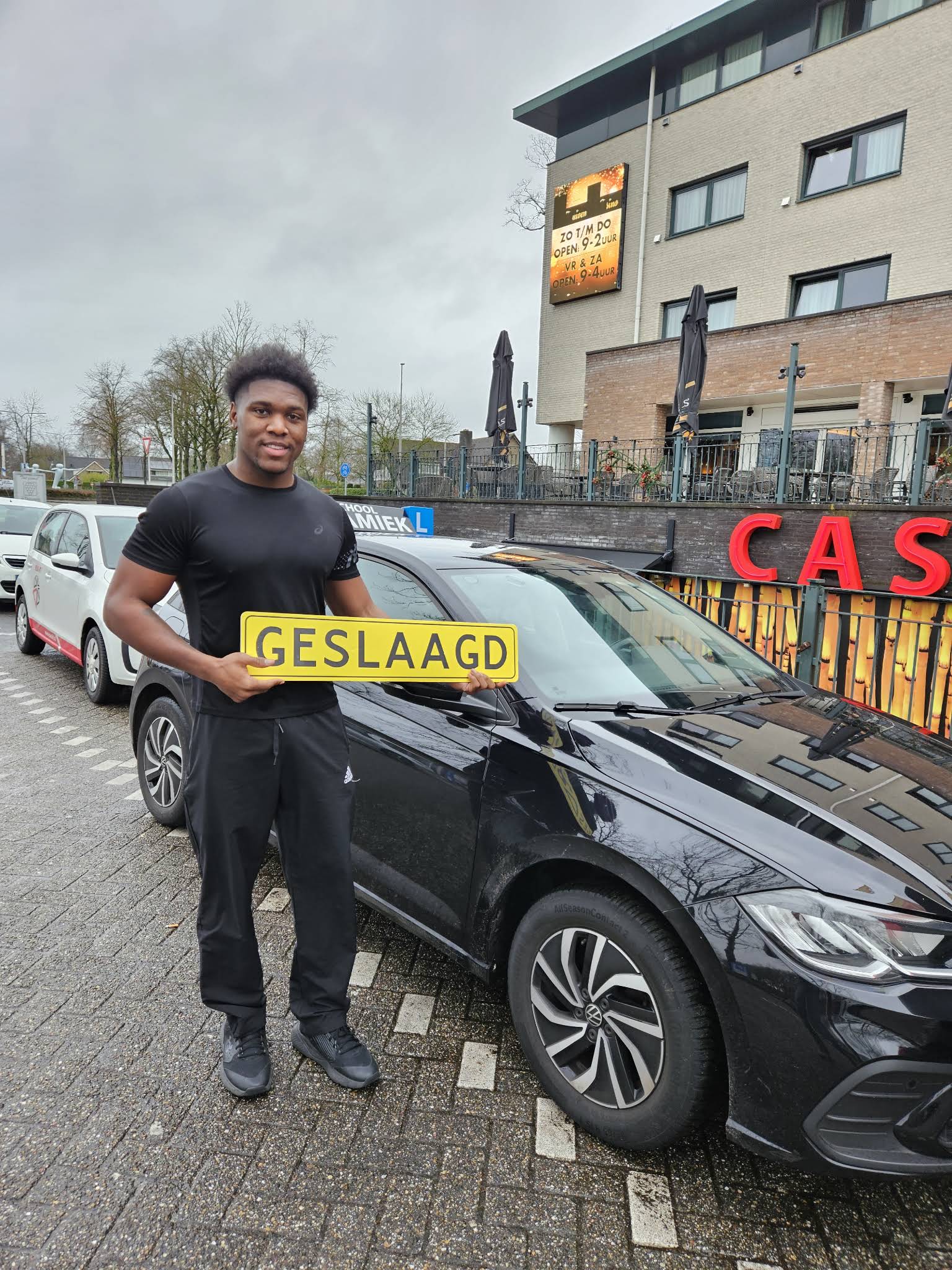 Een trotse leerling staat met een geslaagd-bord voor een lesauto